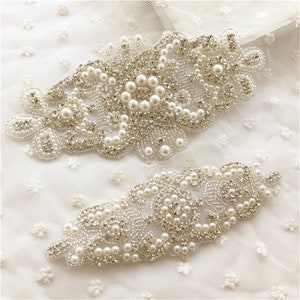 Puede incluir: Dos delicados adornos plateados con detalles de perlas. Los adornos tienen forma de diseños florales y son perfectos para añadir un toque de elegancia a la ropa o las manualidades.