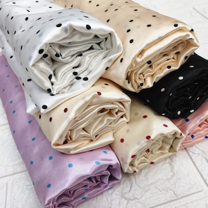 May include: Rolled fabric bolts in various colors, including white, cream, black, and lavender. Each fabric features a polka dot pattern in contrasting colors, such as black, red, and blue. The fabric has a satin sheen.