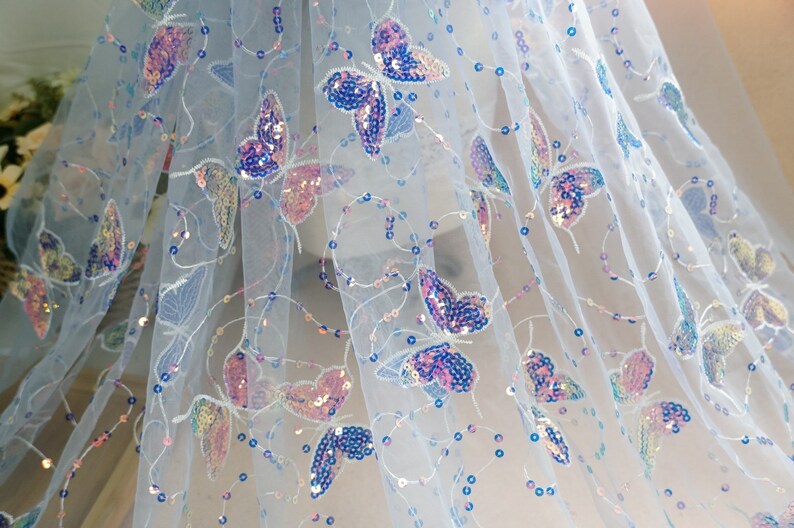 May include: White sheer fabric with iridescent butterfly sequins. The fabric has a delicate, airy feel and is perfect for adding a touch of sparkle to any project.