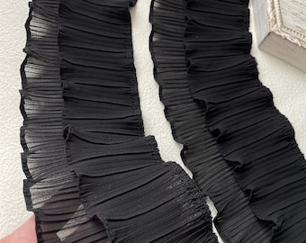 May include: Close-up of two strips of black pleated fabric. The fabric has a delicate, sheer appearance with a ruffled edge. The pleats run horizontally across the strips, creating a textured effect. The fabric is likely used for decorative purposes.
