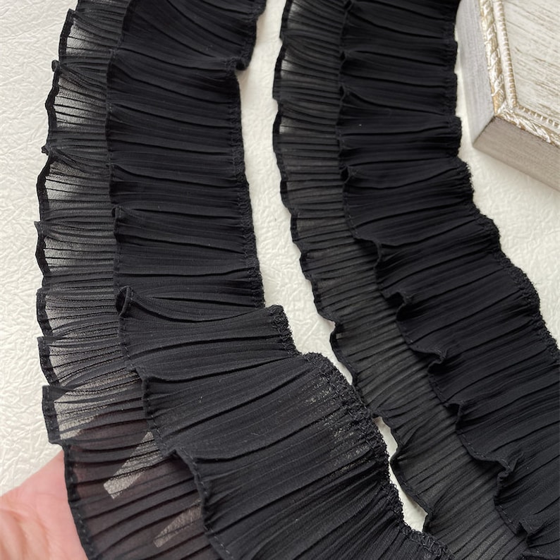 May include: Close-up of two strips of black pleated fabric. The fabric has a delicate, sheer appearance with a ruffled edge. The pleats run horizontally across the strips, creating a textured effect. The fabric is likely used for decorative purposes.