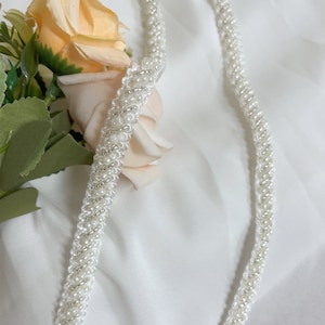 May include: White beaded chain with a braided design. The chain is made of small, white beads that are woven together to create a rope-like texture.