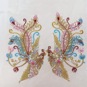 May include: Two embroidered phoenix appliques with gold, pink, blue, and red sequins and beads. The phoenixes have detailed feather patterns and are set against a white background. The design includes floral and swirling elements.