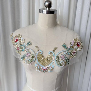 May include: An ornate, embroidered applique with a floral design. The applique features gold, red, pink, and teal sequins and beads. The design is on a sheer, white mesh backing, and is displayed on a mannequin.