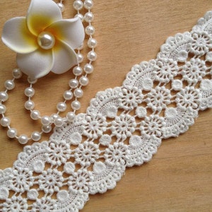 May include: White crochet lace trim with a delicate floral pattern. The lace is shown with a pearl necklace and a white flower.