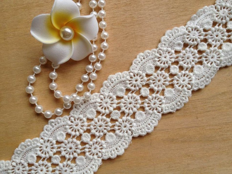 May include: White crochet lace trim with a delicate floral pattern. The lace is shown with a pearl necklace and a white flower.
