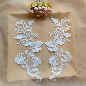 May include: Two symmetrical white lace appliqués featuring intricate floral and leafy embroidery patterns. The lace pieces are positioned side by side on a sheer beige fabric, with a small cluster of artificial pink and yellow flowers above.
