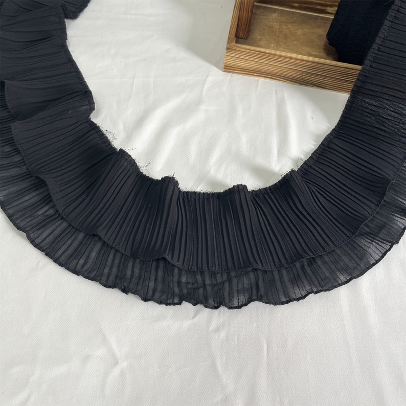 May include: Black pleated fabric trim with a ruffled edge. The trim is made of a lightweight material and is arranged in a curved shape against a white background. The fabric is likely used for sewing, crafting, or adding detail to clothing.