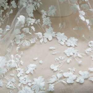 May include: White floral lace fabric featuring a delicate pattern of flowers and leaves. The lace is sheer and has a soft, flowing texture.