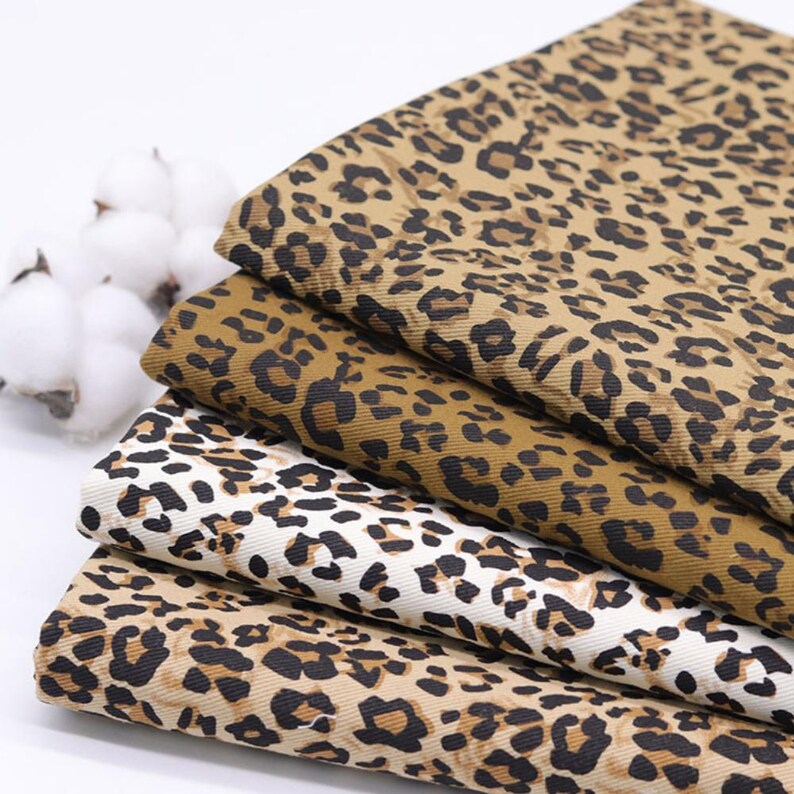 May include: Close-up of folded fabric with a leopard print pattern. The fabric is in shades of brown, tan, and white, with black spots. The fabric is stacked in a neat pile. Cotton buds are in the background.