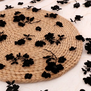 以下が含まれることがあります： ベージュのメッシュ生地に黒い花の刺繍が施されています。生地は、丸い織りのマットの上にかけられています。