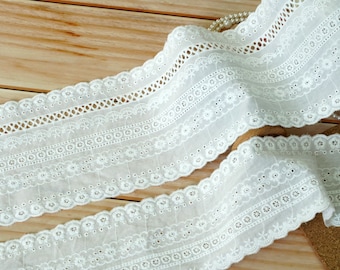 May include: White cotton lace trim with a scalloped edge and delicate floral embroidery. The lace has a repeating pattern of small flowers and circles.