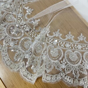 Pu&ograve; includere: Tessuto di tulle bianco con un bordo di perline e paillettes. Il bordo presenta un bordo smerlato e un motivo floreale.