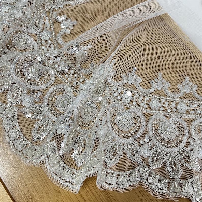 Pu&ograve; includere: Tessuto di tulle bianco con un bordo di perline e paillettes. Il bordo presenta un bordo smerlato e un motivo floreale.
