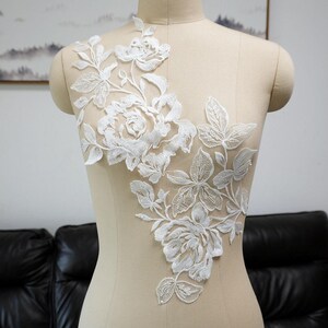May include: White floral lace applique featuring detailed embroidered roses and leaves. The delicate design is displayed on a mannequin, showcasing the intricate patterns and textures of the lace. Ideal for bridal wear or craft projects.