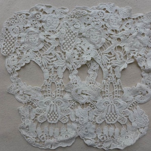 May include: Two white lace skull shapes, with intricate floral designs, are placed on a beige fabric background.