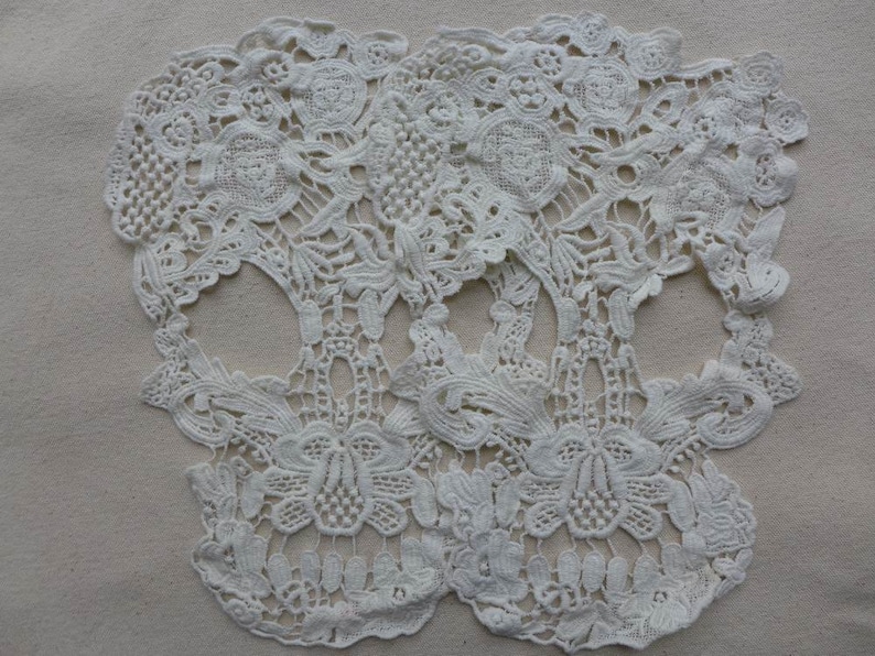 May include: Two white lace skull shapes, with intricate floral designs, are placed on a beige fabric background.