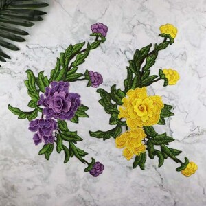 May include: Two embroidered floral patches. One patch features purple flowers and buds with green leaves. The other patch has yellow flowers and buds with green leaves. The patches are on a white and gray marble background.