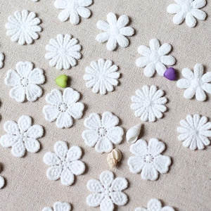 May include: A collection of white, crocheted flower appliques scattered on a beige fabric surface. The flowers vary in style, with some having rounded petals and others with pointed ones. Small decorative elements, including a green heart, a purple heart, and seashells, are also present.