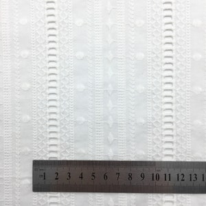 May include: White cotton fabric with an embroidered pattern of small circles and geometric shapes. A ruler is placed on the fabric, showing measurements in inches and centimeters.