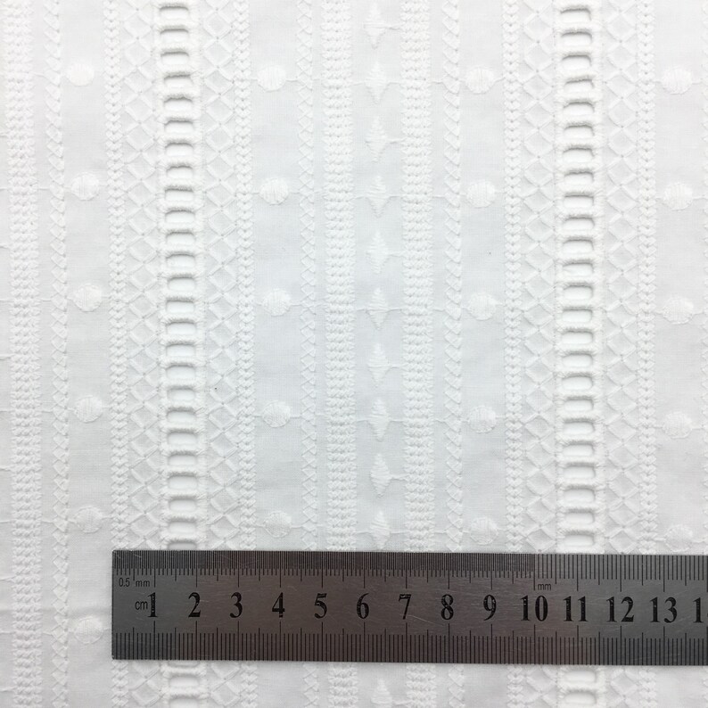 May include: White cotton fabric with an embroidered pattern of small circles and geometric shapes. A ruler is placed on the fabric, showing measurements in inches and centimeters.