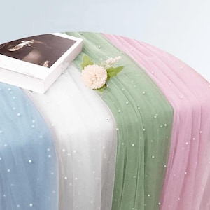 May include: A collection of sheer tulle fabric runners in blue, white, sage green, and soft pink, each embellished with scattered white faux pearls. A white rectangular box and a single artificial flower are placed on the fabric layers.