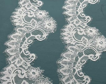 May include: Close-up of two strips of white lace with a floral design. The lace features intricate patterns of flowers, leaves, and delicate openwork, with a scalloped edge. The lace is set against a teal background.