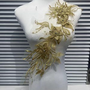 May include: A gold embroidered lace applique featuring a floral and vine motif with intricate stitching and small clear beads. The design is arranged in an asymmetrical, elongated shape, showcasing detailed petals and leaves on a sheer mesh base.