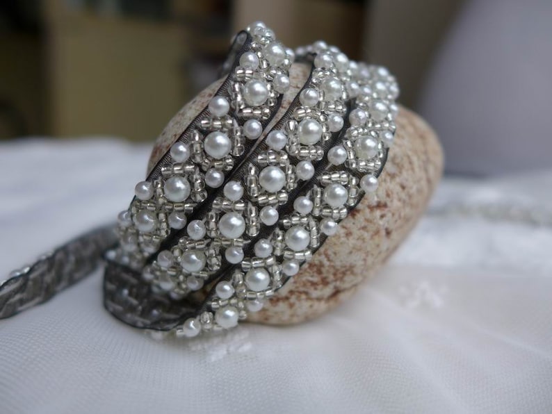 May include: Three strands of black ribbon trim with white pearl beads and silver accents. The trim is arranged in a circle on a light brown rock.