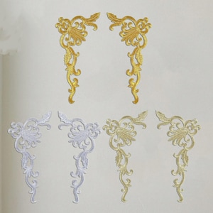May include: Embroidered corner appliques in gold and white. The appliques feature a floral and scroll design. The gold appliques are in the top row, and the white appliques are in the bottom row. The appliques are arranged on a white background.