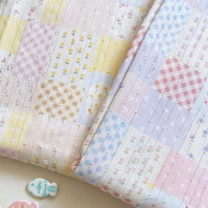 May include: A textured fabric featuring a patchwork design of pastel squares in yellow, pink, blue, and lavender. The pattern includes small floral prints, gingham checks, and diamond motifs, all accented with a subtle raised dot texture throughout.