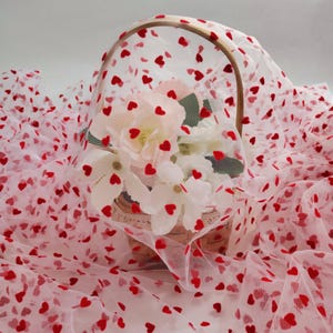 May include: White tulle fabric scattered with small red hearts, covering a basket filled with white flowers. The basket has a wooden handle. The fabric is arranged in soft folds, creating a romantic and delicate appearance.
