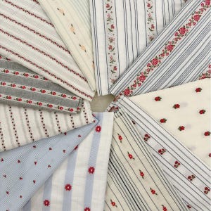 May include: A collection of folded fabric swatches featuring various patterns including thin blue, grey, and white stripes, small red floral embroidery, and delicate rosebud motifs. The textiles display a mix of solid colours and striped designs.