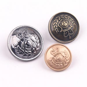 Pode incluir: Três botões redondos de metal em prata, bronze e ouro. Cada botão apresenta um emblema detalhado com uma coroa, folhas de louro e um desenho central. Os botões são provavelmente para roupas ou decoração.