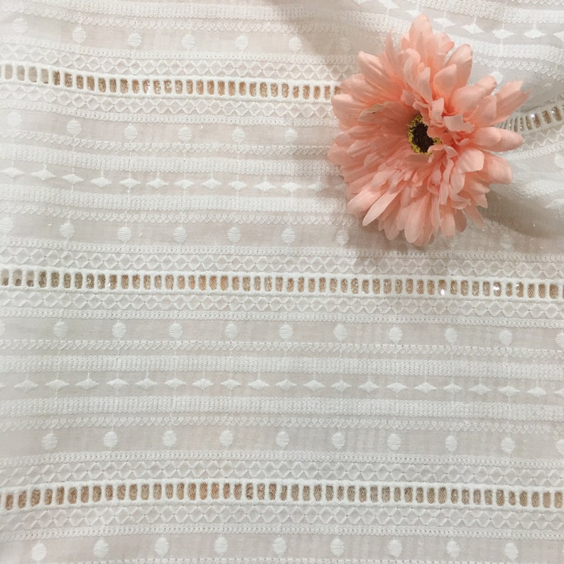 May include: White cotton fabric with a delicate embroidered pattern of circles, diamonds, and lines. A single pink flower is placed on the fabric.