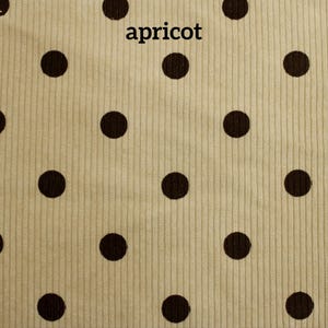 May include: A light beige corduroy fabric featuring a repeating pattern of dark brown polka dots. The word apricot is printed in black lowercase letters near the top center of the textile, which has a distinct vertical ribbed texture.