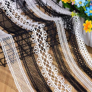 May include: Assortment of white and black lace ribbons with various floral and geometric patterns. The ribbons are displayed on a black wire rack, showcasing the different designs and textures. Decorative craft supplies.
