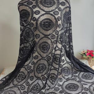 Peut inclure: Un tissu en dentelle noire pr&eacute;sentant un motif r&eacute;p&eacute;titif de grands cercles avec des centres floraux complexes et des d&eacute;tails en maille. Le tissu est drap&eacute; sur un mannequin, mettant en valeur la texture semi-transparente et le design g&eacute;om&eacute;trique.
