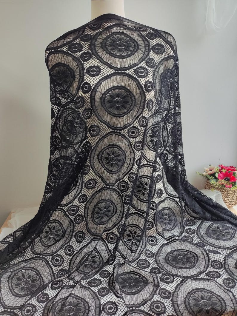 Peut inclure: Un tissu en dentelle noire pr&eacute;sentant un motif r&eacute;p&eacute;titif de grands cercles avec des centres floraux complexes et des d&eacute;tails en maille. Le tissu est drap&eacute; sur un mannequin, mettant en valeur la texture semi-transparente et le design g&eacute;om&eacute;trique.
