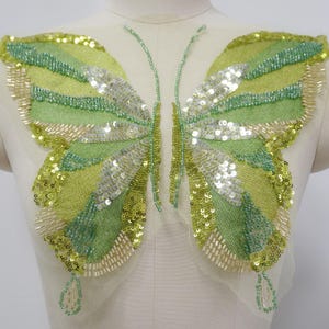 May include: A decorative butterfly applique featuring green, yellow, and silver sequins and beads. The butterfly design has two large wings with a gradient of colours, and a beaded body. The applique is on a sheer, white mesh backing.
