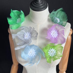 Può includere: Una collezione di cinque spille decorative a forma di fiore di piuma fissate su un manichino. Ogni spilla presenta piume morbide e stratificate nelle tonalità verde brillante, grigio chiaro, lavanda, azzurro e verde lime, con un fiore bianco.