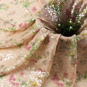May include: Close-up of a beige fabric with a floral pattern and sequins. The fabric features a delicate design with pink, yellow, and green flowers. The sequins add a shimmering effect, creating a luxurious look for the material.