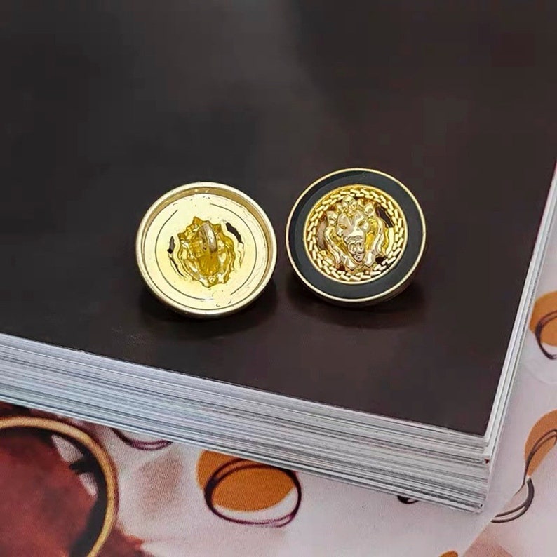 Puede incluir: Dos botones de color dorado con un dise&ntilde;o de cabeza de le&oacute;n. Un bot&oacute;n tiene un fondo negro y el otro tiene un fondo dorado.