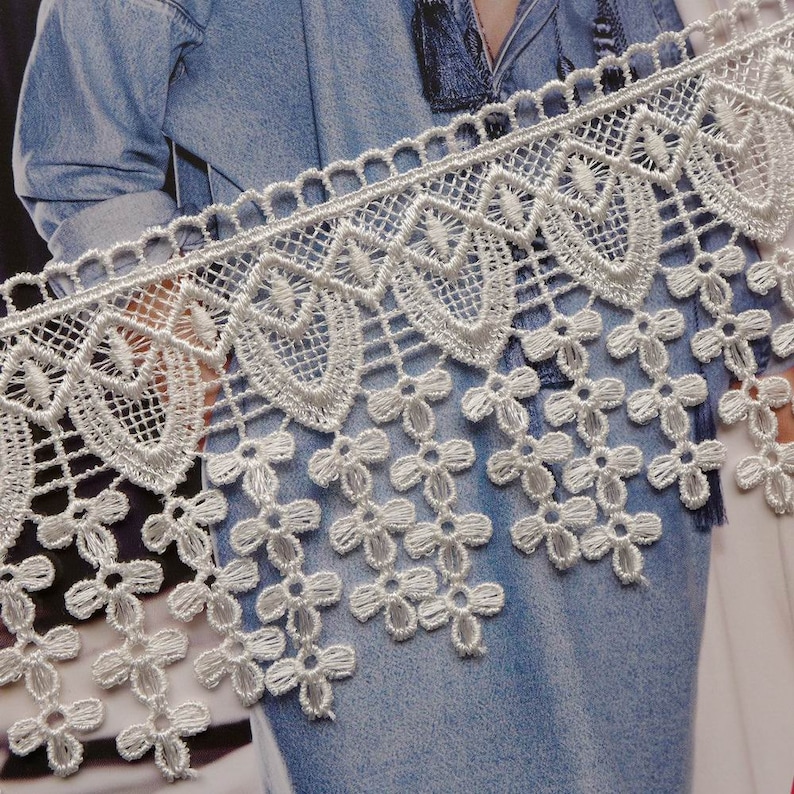 May include: White lace trim featuring a scalloped edge and a fringe of small flowers. The lace is delicate and intricate, and would be ideal for adding a touch of elegance to any project.