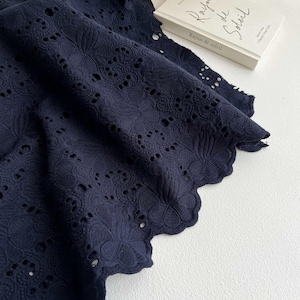 Puede incluir: Primer plano de tela calada azul marino con borde festoneado. La tela está intrincadamente detallada con recortes florales y geométricos. Un libro titulado "Rayon de Soleil" es visible en el fondo.