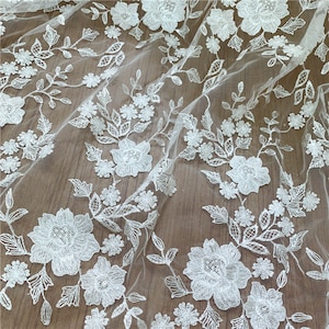 Puede incluir: Primer plano de tela de encaje floral blanco. El encaje presenta flores, hojas y vides bordadas con detalle. El dise&ntilde;o delicado se establece sobre un fondo transparente, creando una apariencia texturizada y elegante. Ideal para ropa de novia o proyectos decorativos.