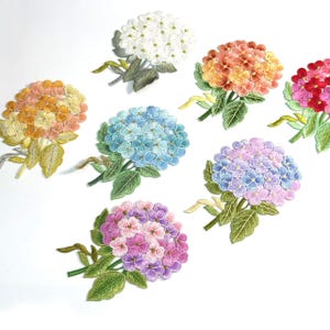 May include: A collection of seven embroidered hydrangea flower patches in various colors including white, orange, yellow, blue, pink, and purple, each featuring detailed green leaves and stems arranged in a scattered pattern on a white background.