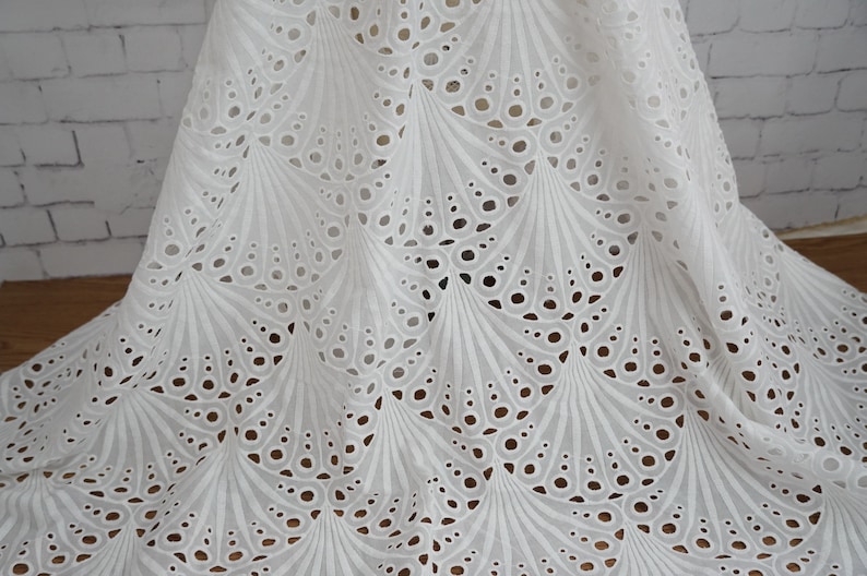 Puede incluir: Tela de algod&oacute;n blanca con un delicado y complejo patr&oacute;n de ojales. El patr&oacute;n presenta un dise&ntilde;o repetitivo de c&iacute;rculos y arcos superpuestos, creando un aspecto fluido y elegante.