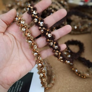 May include: Close-up of two beaded trims in shades of brown and gold. The trims are embellished with small beads and larger, iridescent sequins. The texture of the trim appears soft and fuzzy, with a decorative design.