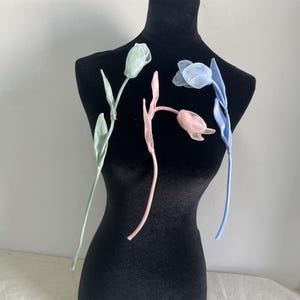 May include: Three artificial flower stems with delicate fabric petals. The stems are green, pink, and blue. The flowers are arranged on a black mannequin.